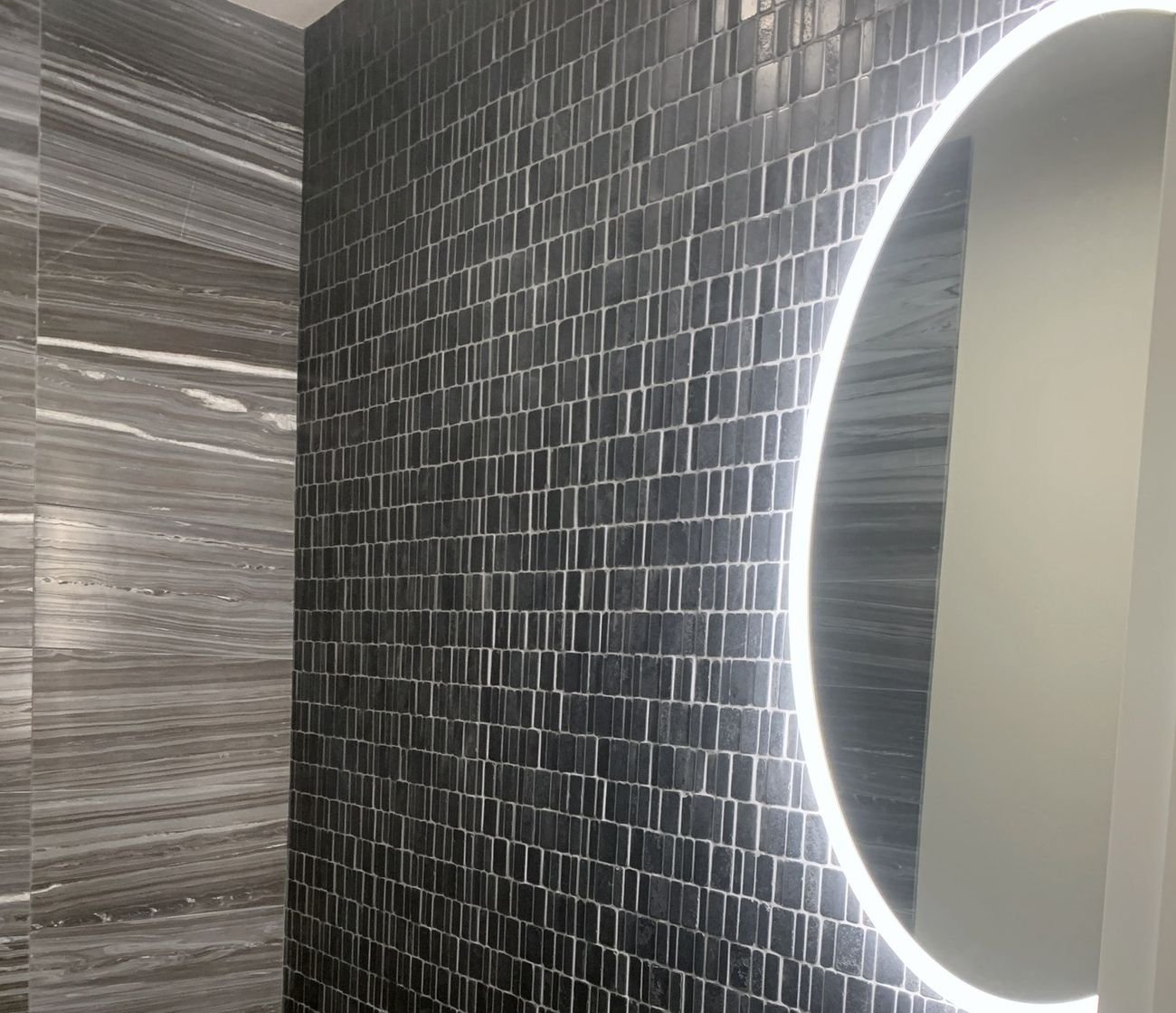 Dark mosaic wall tile beside an illuminated circular mirror
